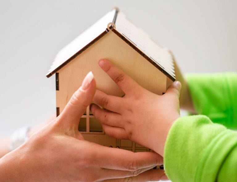 Close-up of mother and child holding a small wooden model house, symbolizing family and home.